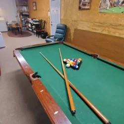 A pool table with two cues, a racked set of balls, and a cue ball. Nearby are chairs, a desk, and wall decorations.