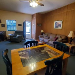 A cozy living room with a wooden dining table, chairs, couch, armchair, ceiling fan, and wall art. Door is slightly open.