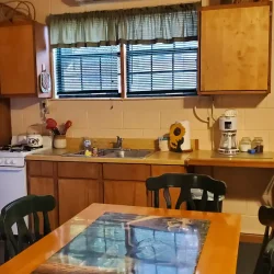 A small kitchen with a table, chairs, refrigerator, stove, sink, coffee maker, and wall clock. Sunflower decor on the counter.