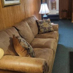 A cozy living room with a brown sofa, patterned cushions, a lit lamp on a side table, and framed artwork on wood-paneled walls.
